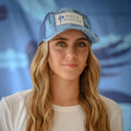 Woman smirking while wearing the Pole Dancer Alpha Patch Camo Trucker Hat in the Water Camo colorway. Standing in front of a blue backdrop.