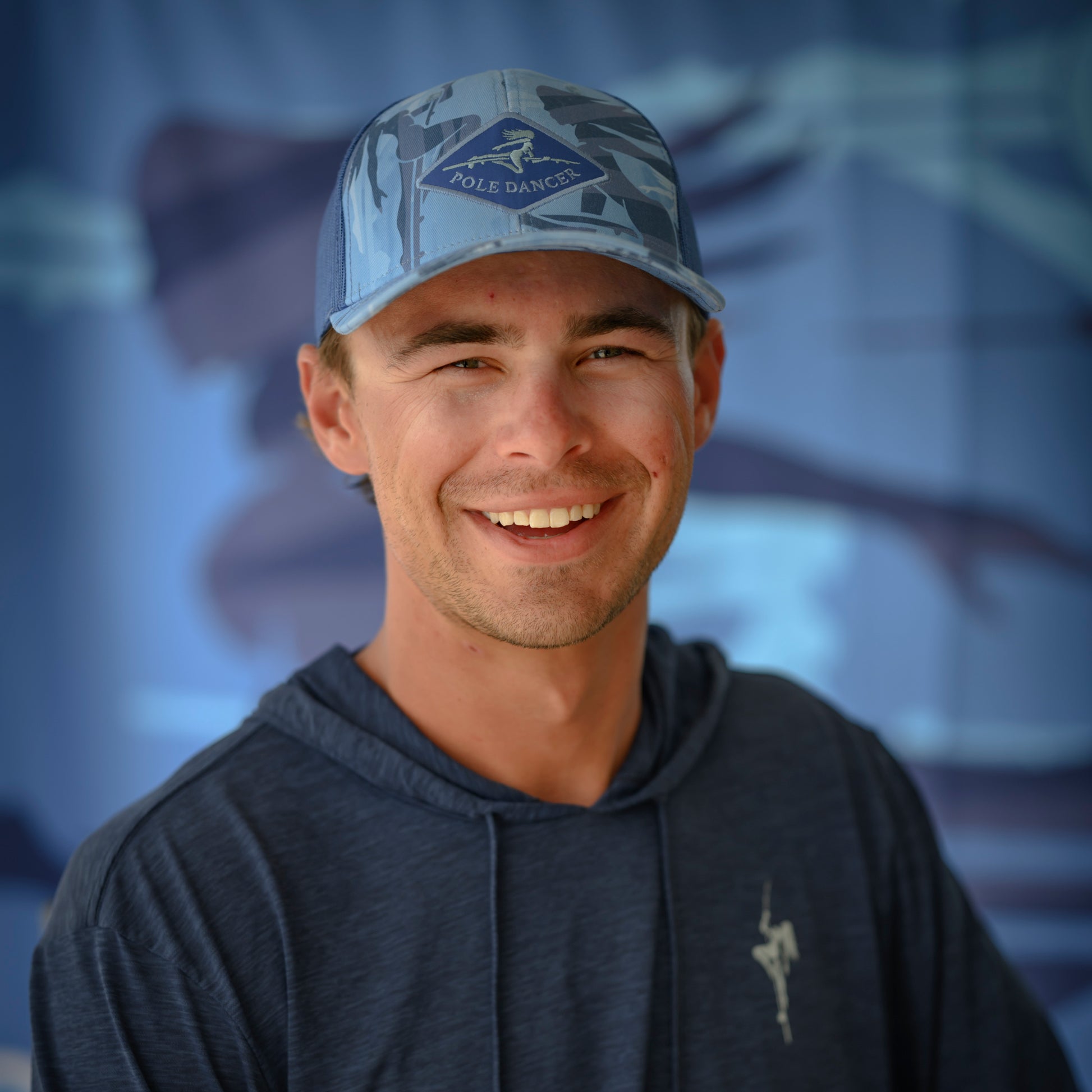 Man wearing the water camo Diamond Patch Camo Trucker Hat and navy performance hoodie in front of a blue backdrop.