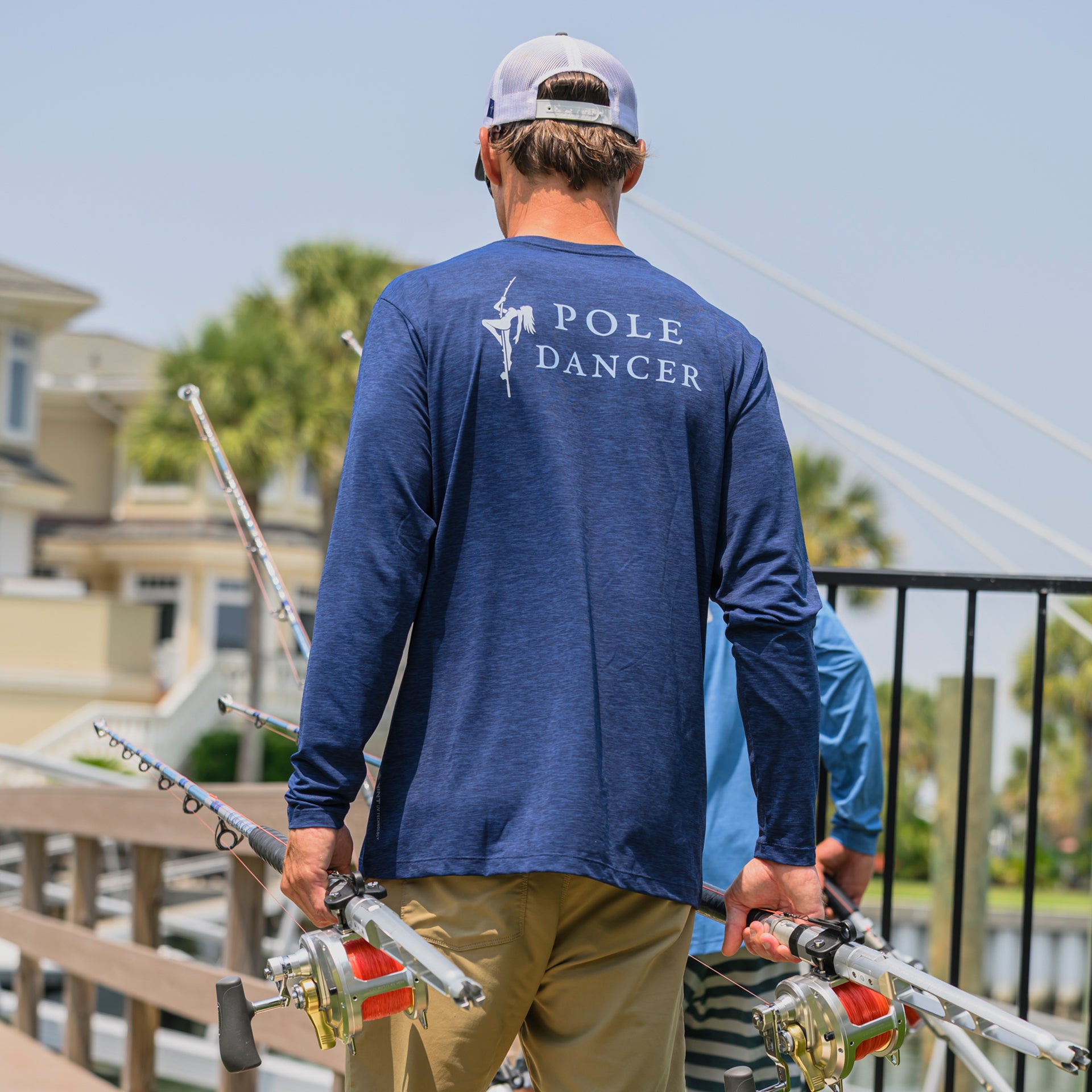 Man wearing the Navy/Heather Pole Dancer Alpha Long Sleeve Performance shirt while walking on a dock carrying several fishing poles.